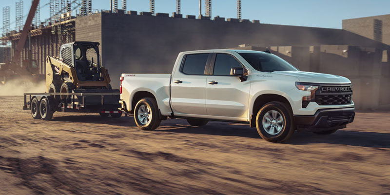 2026 Chevrolet Silverado 1500 white pickup truck towing a small loader through sandy terrain behind a factory, highlighting its power, durability, and work-ready capability.